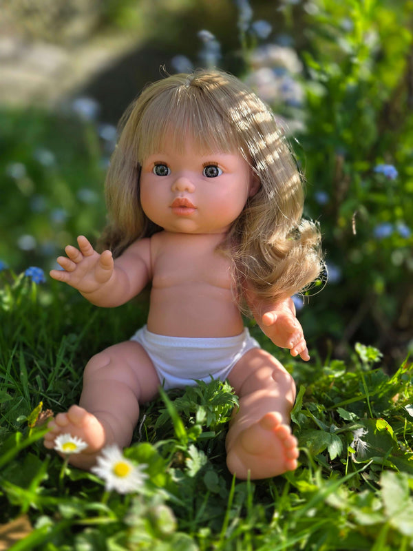 Mini Colettos doll Kate with blonde hair and blue eyes sitting in grass among flowers.