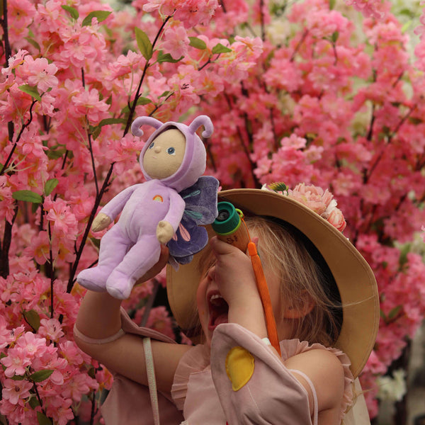 Child joyfully holding Olli Ella Dinky Dinkum Doll Betty Butterfly, surrounded by beautiful pink blossoms.