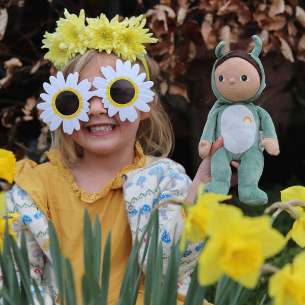 Child in flower crown and oversized sunglasses holding Gideon Grasshopper doll among blooming daffodils.