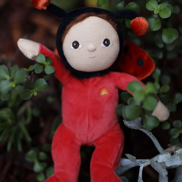 Lola Ladybug doll wearing red suit and black hood, surrounded by greenery, ready for adventures.