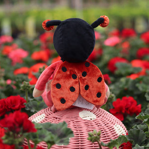 Lola Ladybug plush doll with spotty wings sitting on a pink basket surrounded by blooming red flowers.