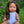 Mini Colettos, Isabel (Hispanic Girl, Brown Straight Hair and Blue Eyes) sitting outdoors in a vibrant garden.
